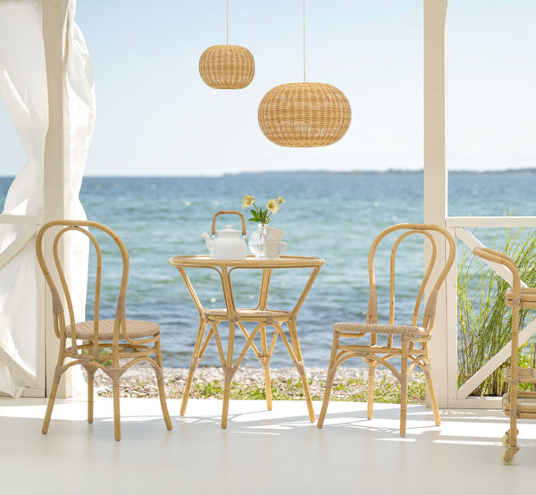 Dies ist ein Lifestyle-Bild von Tony Außen-Cafétisch in natural aluminium artfibre. Auf einer Strandterrasse mit Stühlen, Pendelleuchte und Meerblick.