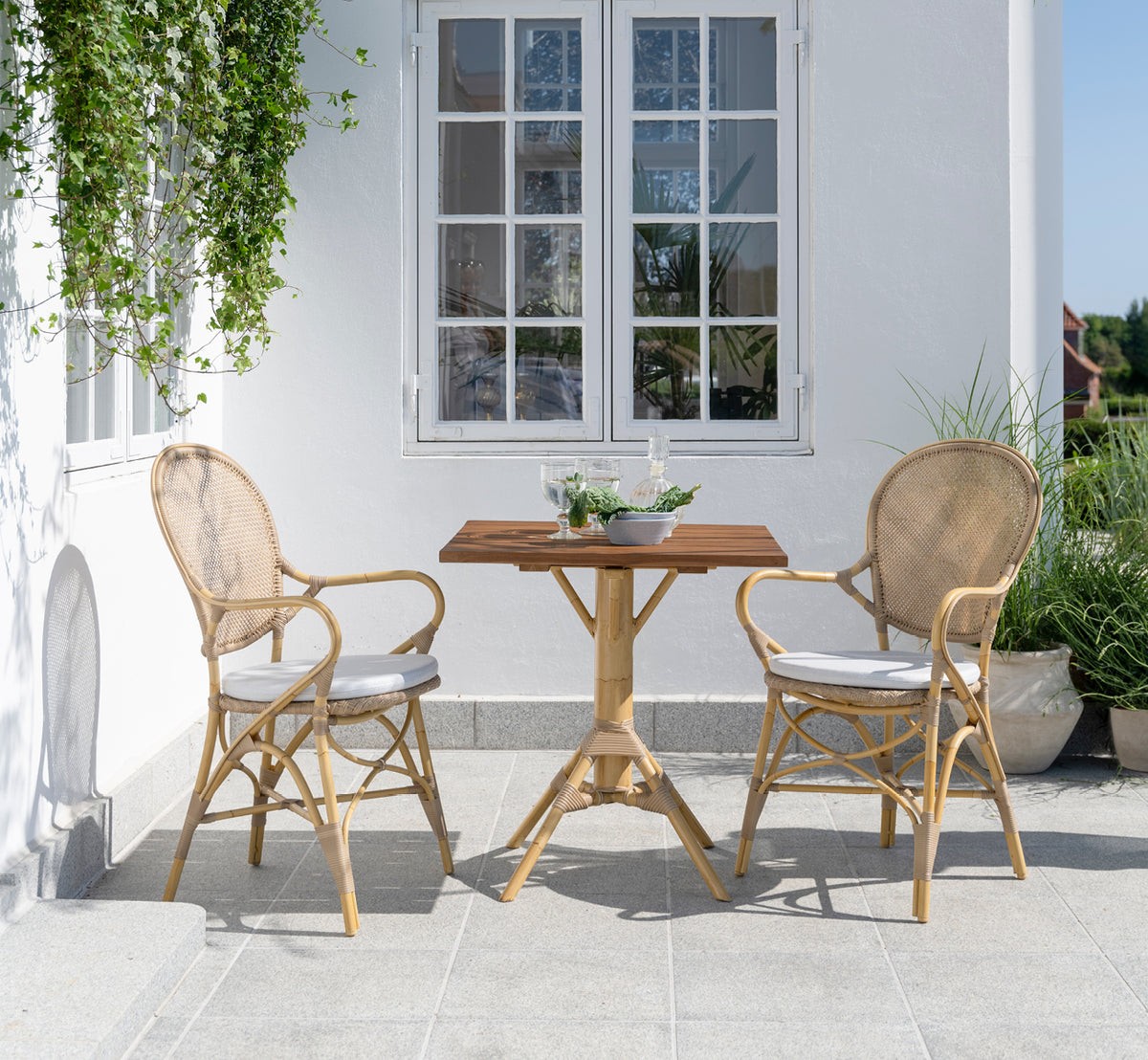 Dies ist ein Lifestyle-Bild von Nicole Cafe Table i natural aluminium artfibre. Auf einer Steinterrasse mit Ofelia-Stühlen und Grünpflanzen.