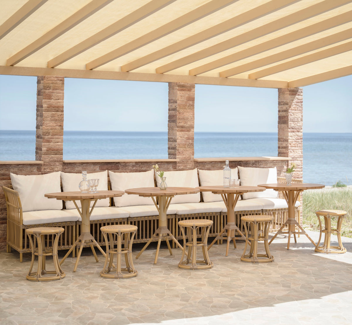 Dies ist ein Lifestyle-Bild von Nicole Cafétisch-Teak Ø80 cm in natural aluminium artfibre. Bank mit Hockern und Cafétischen unter einer Pergola.