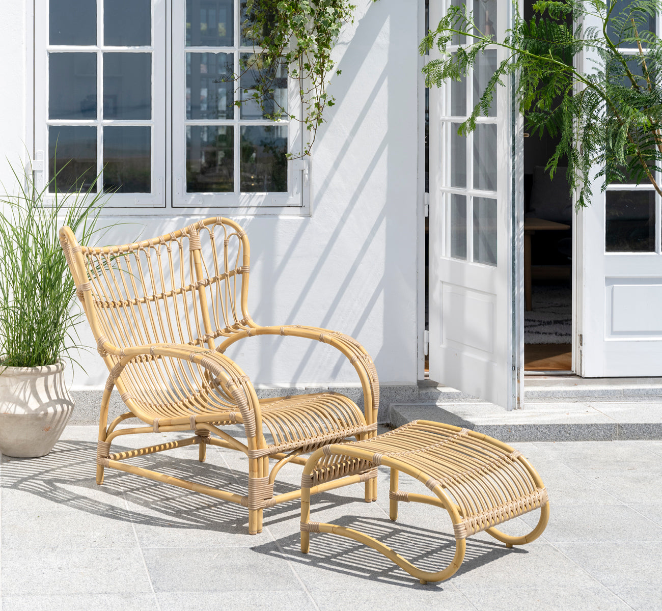 Dies ist ein Lifestyle-Bild von Teddy Außen-Fußhocker in natural aluminium artfibre. Mit dem passenden Loungesessel auf einer sonnigen gepflasterten Terrasse kombiniert.