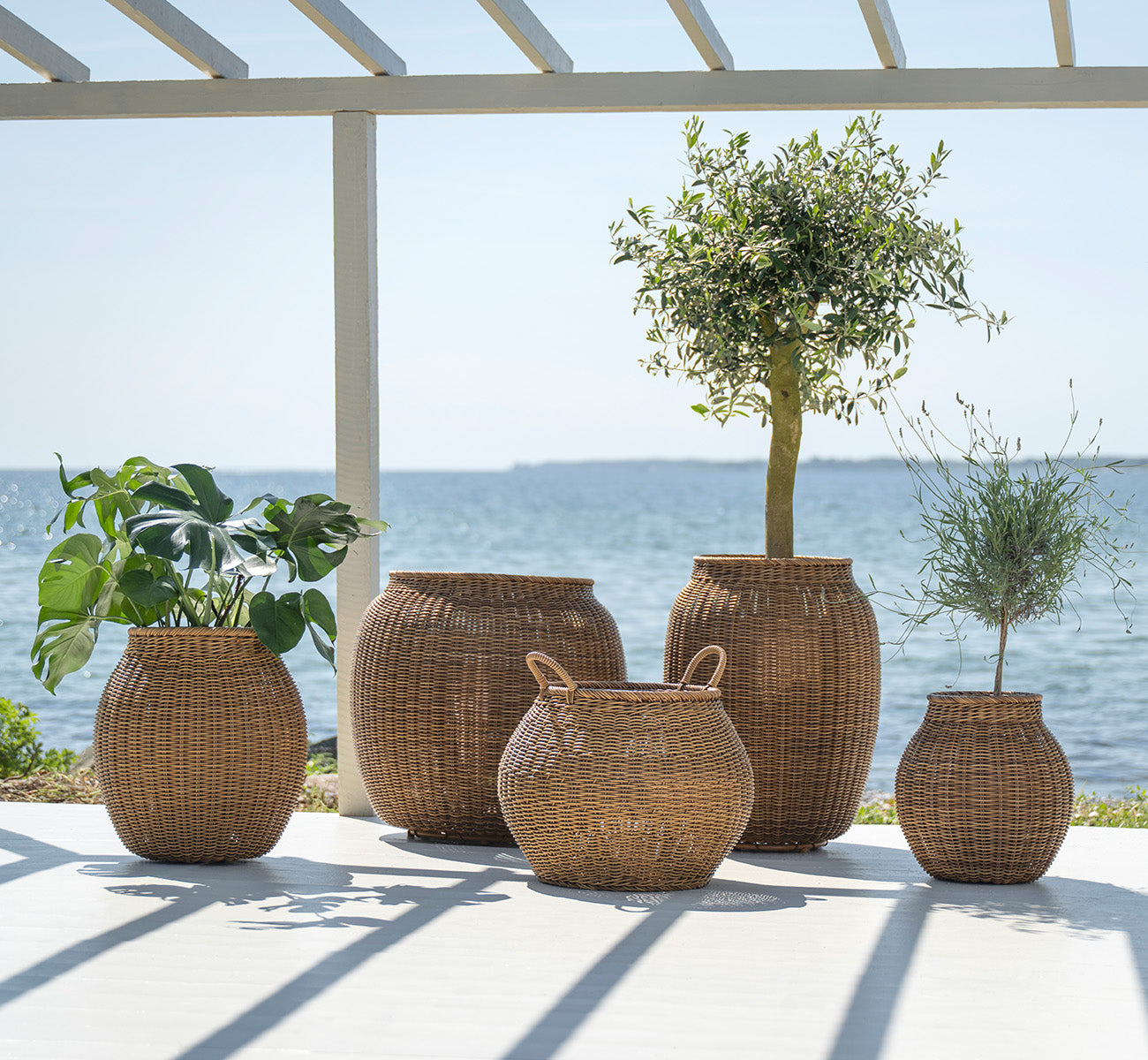 Dies ist ein Lifestyle-Bild von Alma Korb in chestnut core weave ArtFibre Aluminium, im Freien auf einer sonnigen Terrasse mit weißer Mauer und.