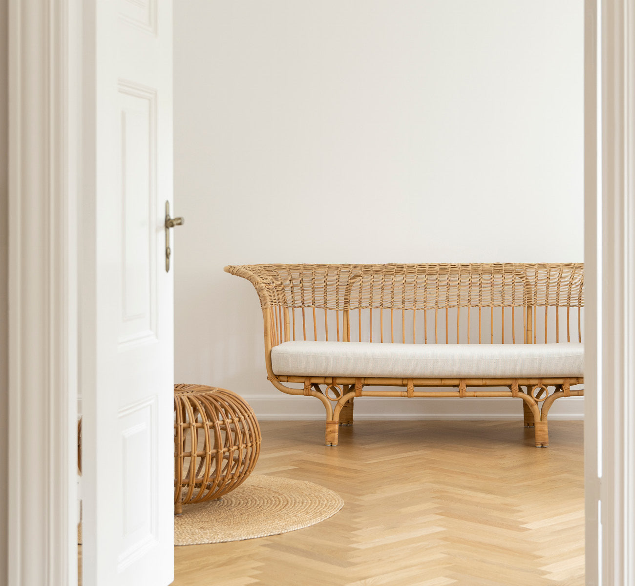 Dies ist ein Lifestyle-Bild von Belladonna Sofa in polished natural rattan, in einem hellen skandinavischen Interieur durch einen weißen Türrahmen.