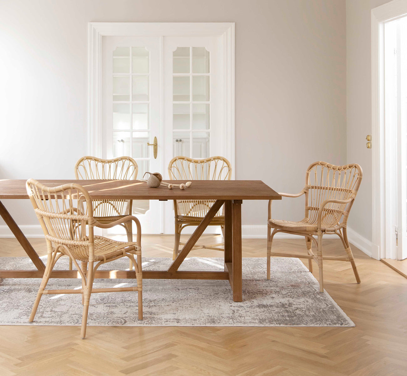 Dies ist ein Lifestyle-Bild von Lucas Teak Dining Table 240X100 cm in teak indoor. Mit Rattanstühlen in einem hellen Raum mit Fischgrät-Eichenboden eingerichtet.
