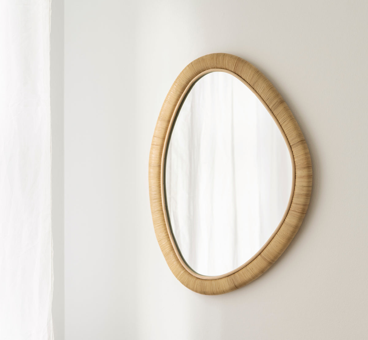 Dies ist ein Lifestyle-Bild von Malou Spiegel in natural rattan. Organische ovale Form an weicher weißer Wand mit Vorhang montiert.