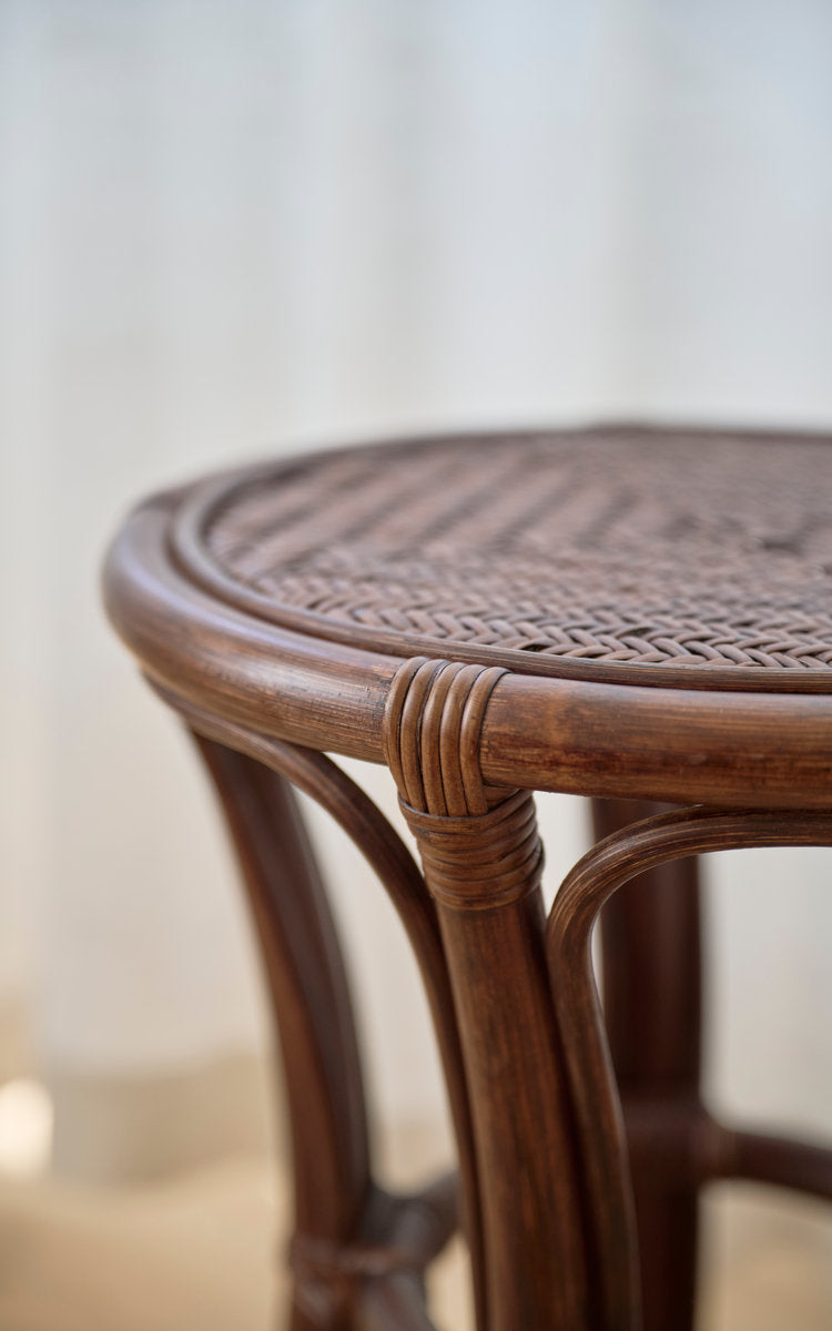 Dies ist ein Nahaufnahmebild von Bella Hocker in brown rattan. Runder geflochtener Sitz und geschwungene Rattanbeine mit Umwicklungen.