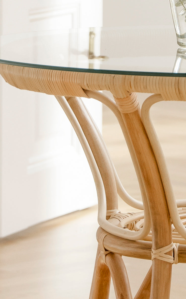 Dies ist ein Nahaufnahmebild von Tony Café Table in natural rattan. Detail der runden Glasplatte mit geflochtener Rattankante und gebogenen Beinen.