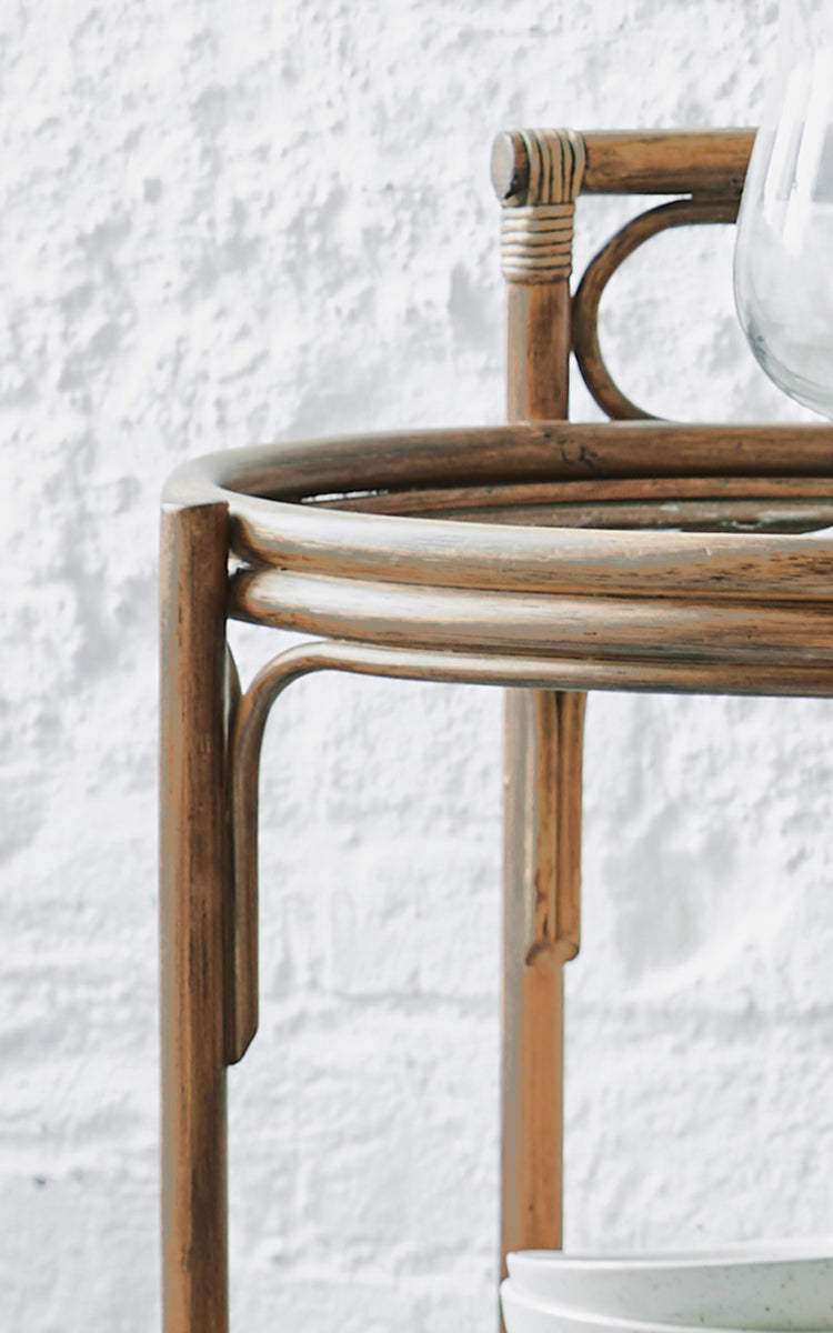 Dies ist ein Nahaufnahmebild von Romeo Servierwagen in antique rattan. Detail des runden Rattan-Servierwagen-Rahmens vor einer weißen Backsteinwand.