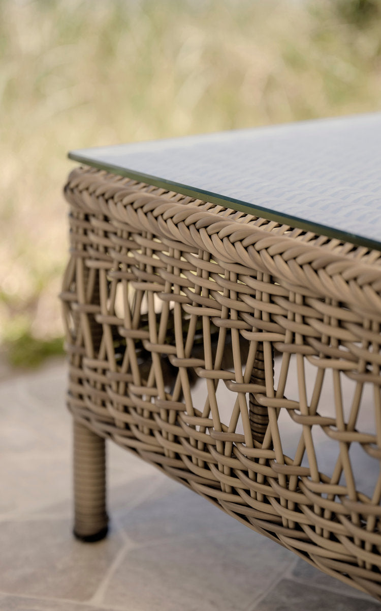 Dies ist ein Nahaufnahmebild von Mona Außen-Modulsofa-Pouf Module w. Glass in antique grey artfibre aluminium. Offenes Geflecht unter einer Glasplatte.