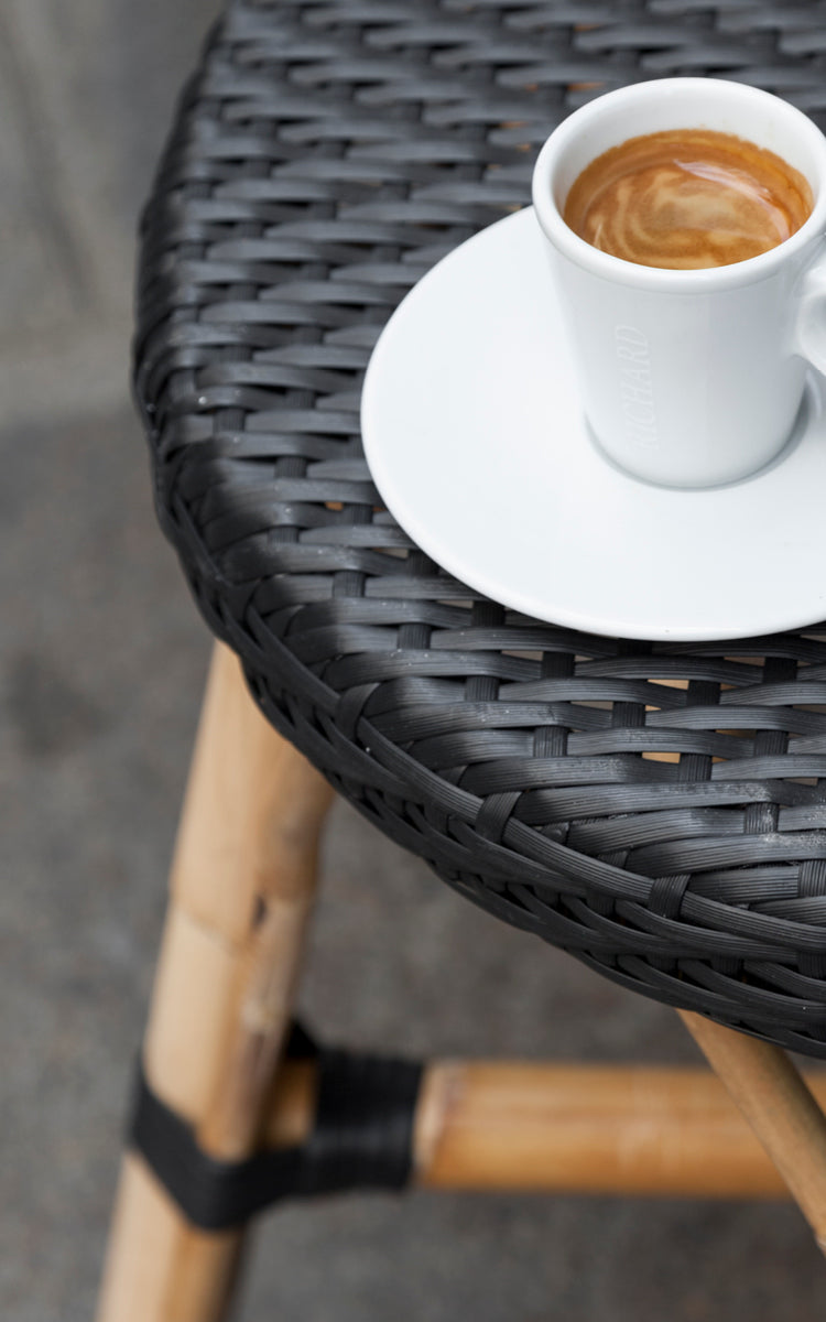 Dies ist ein Nahaufnahmebild von Simone Hocker in black ArtFibre Skin on Rattan frame. Detail der geflochtenen Sitzfläche mit Espressotasse.