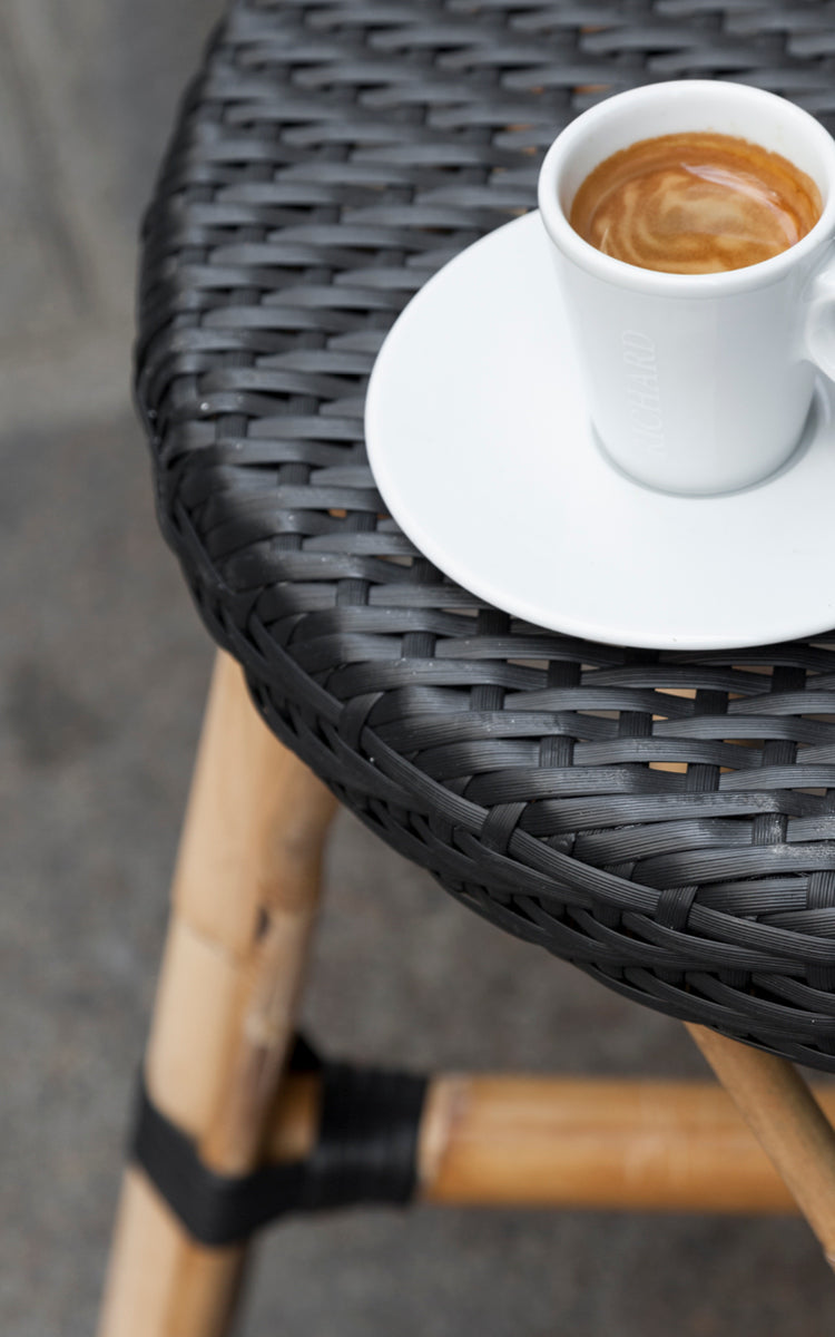 Dies ist ein Nahaufnahmebild von Simone Barstuhl in black ArtFibre Skin on Rattan frame. Detail der geflochtenen Sitzfläche mit Espressotasse.