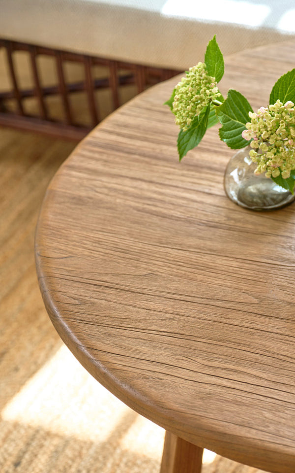 Dies ist ein Nahaufnahmebild von Aksel Couchtisch Ø100 cm in teak indoor. Runde Teakplatte mit Hortensienvase auf einem Juteteppich.