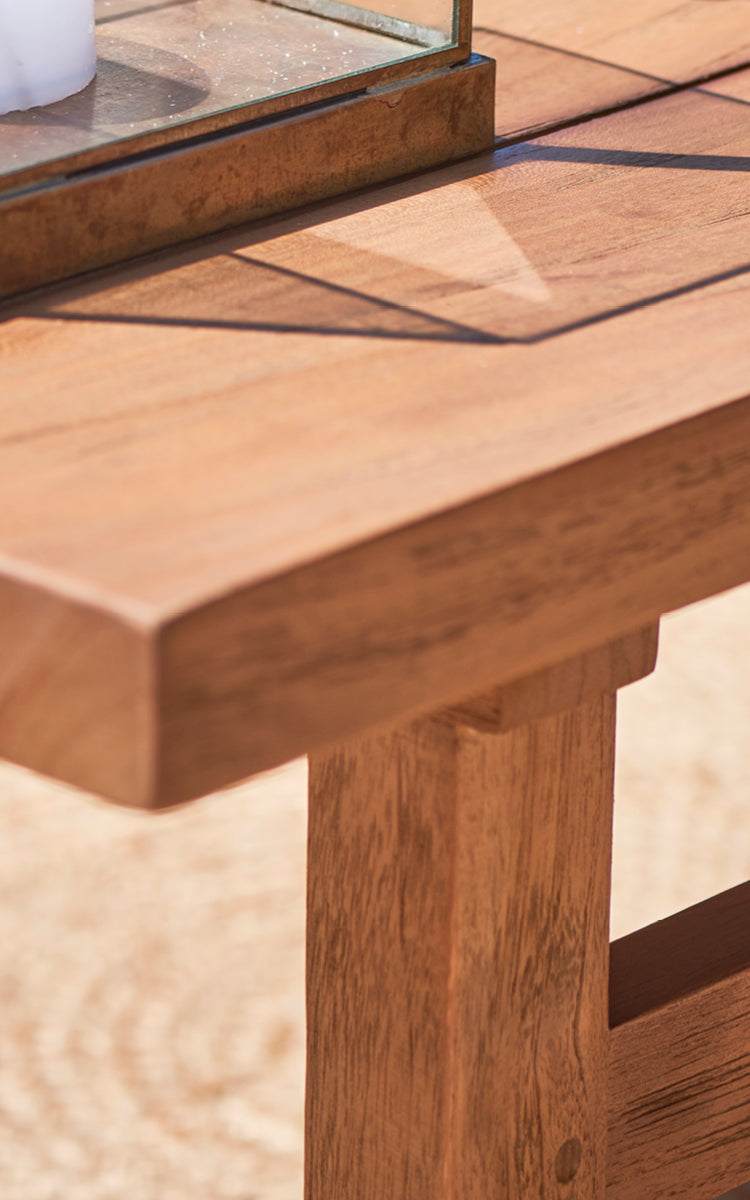 Dies ist ein Nahaufnahmebild von Julian Teak Lounge Table 120x120 cm in teak outdoor. Die massive Teakoberfläche und die dicke Kante sind im warmen Sonnenlicht gezeigt.