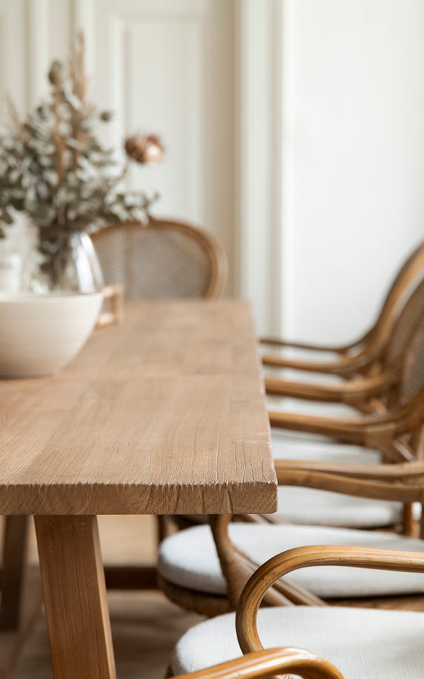 Dies ist ein Nahaufnahmebild von Lucas Teak Dining Table 300X100 cm in teak indoor. Die massive Teakkante ist mit Rattanstühlen im Hintergrund gezeigt.
