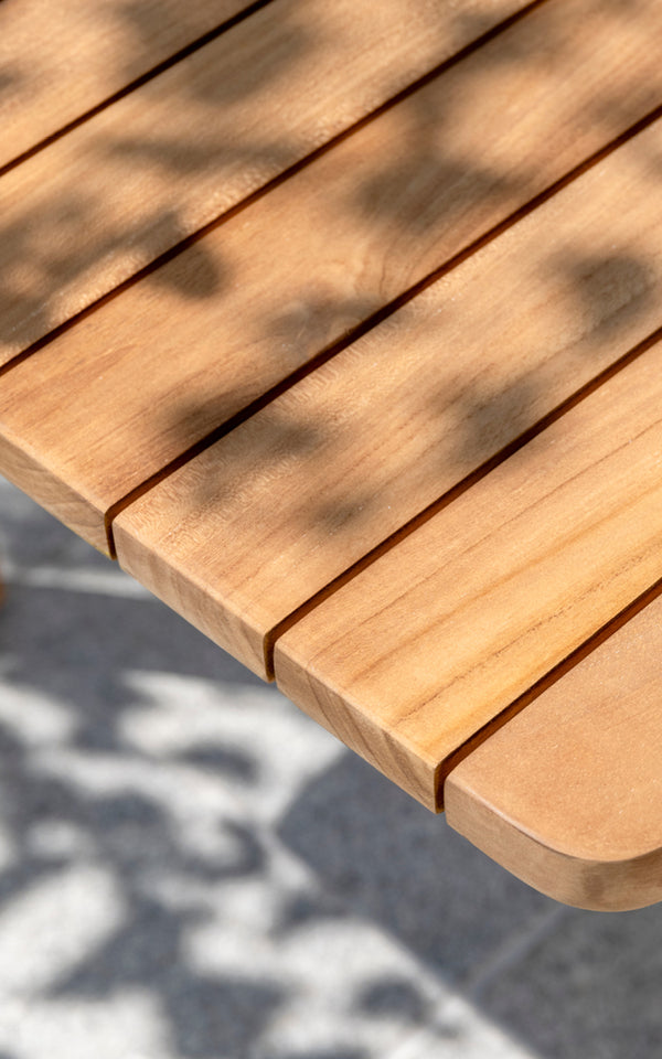 Dies ist ein Nahaufnahmebild von Tobias Exterior Teak Table 205x91 cm in teak outdoor. Detail der Teakbohlen-Tischplattenkante und des gesprenkelten Sonnenlichts.