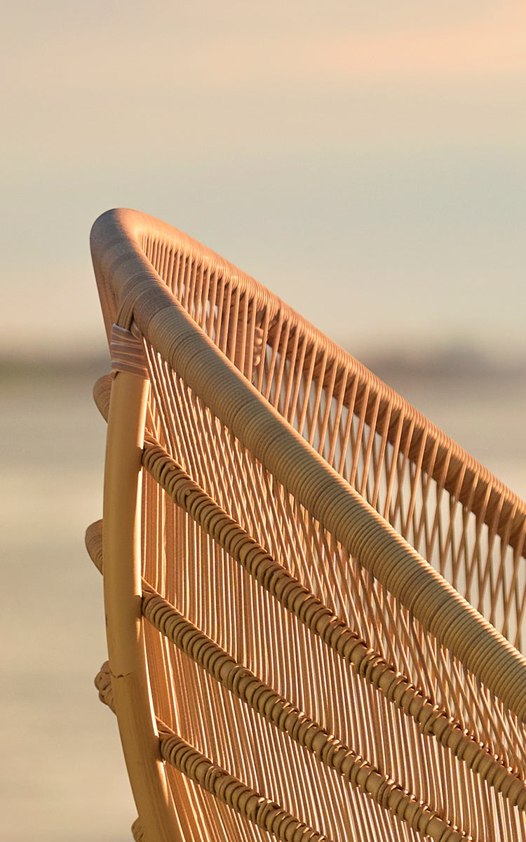 Dies ist ein Nahaufnahmebild von Luna Außen-Lounge-Sessel in natural aluminium artfibre. Die gewebte Rückenlehne leuchtet im goldenen Sonnenuntergangslicht am Meer.