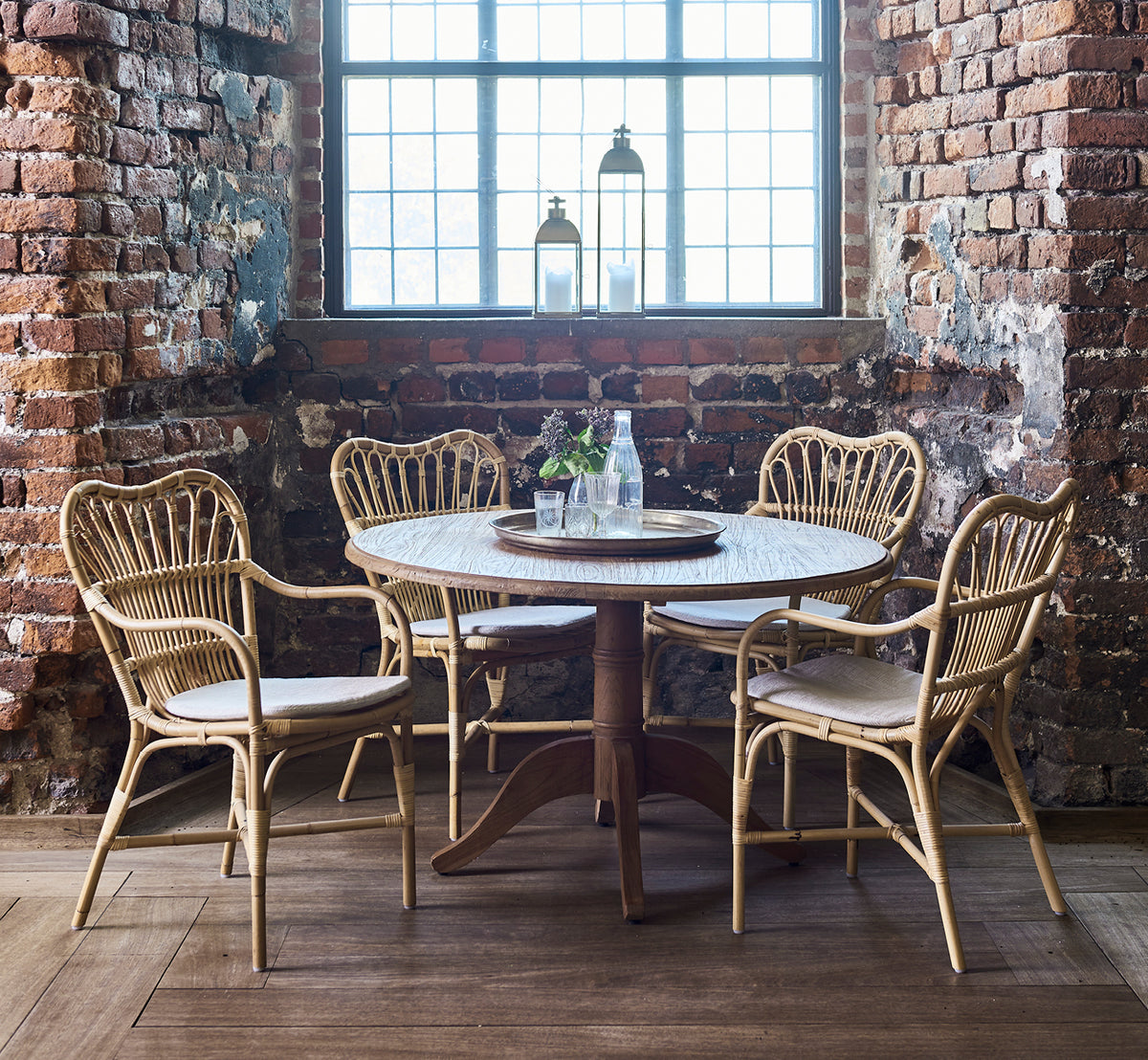 Dies ist ein Lifestyle-Bild von Margret Esszimmerstuhl in natural rattan. Rustikales Backsteininterieur mit rundem Tisch und Laterne auf der Fensterbank.