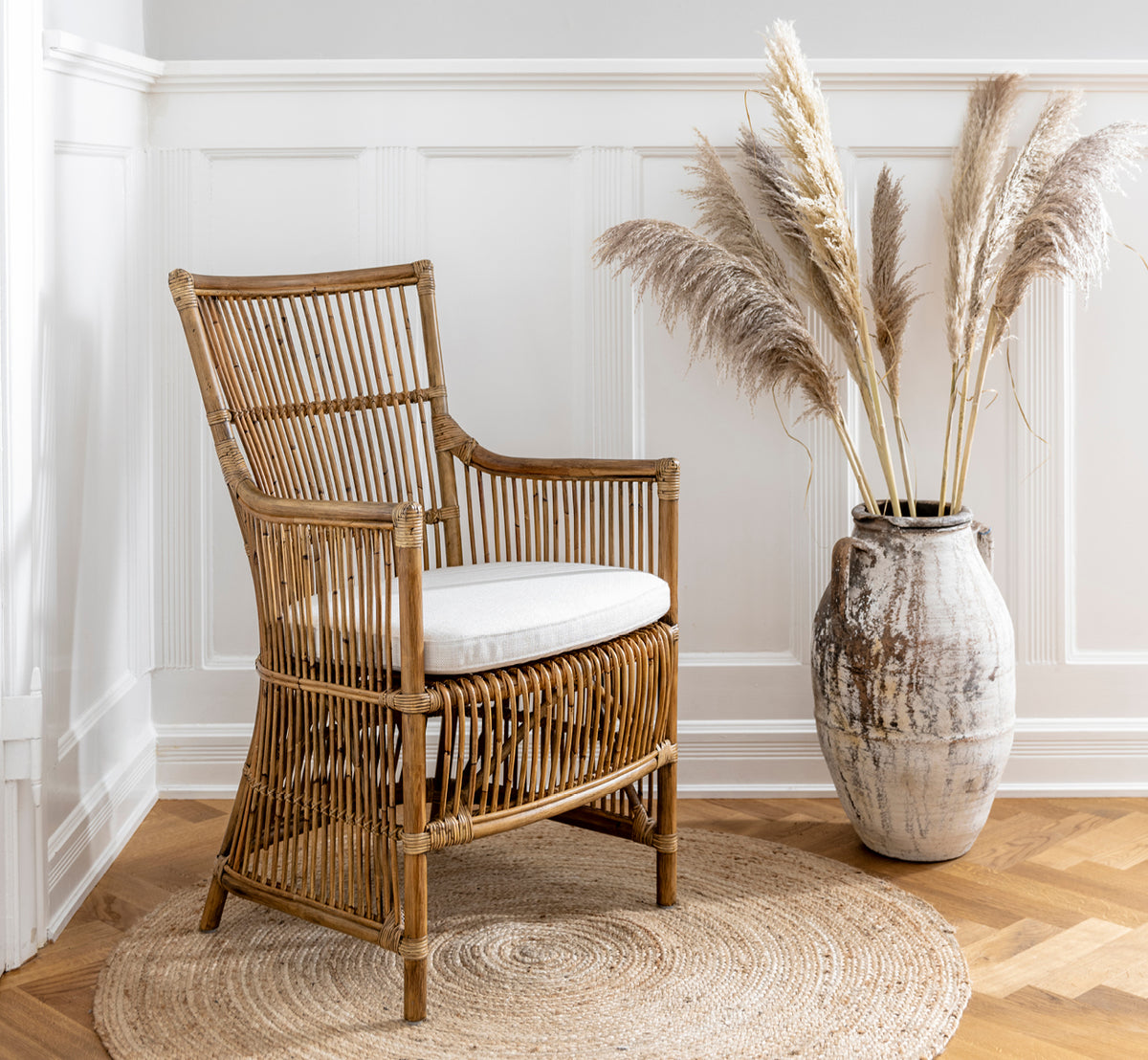 Dies ist ein Lifestyle-Bild von Davinci Esszimmerstuhl in antique rattan, in einem hellen Interieur mit weißen Wandpaneelen und Fischgrätparkett.