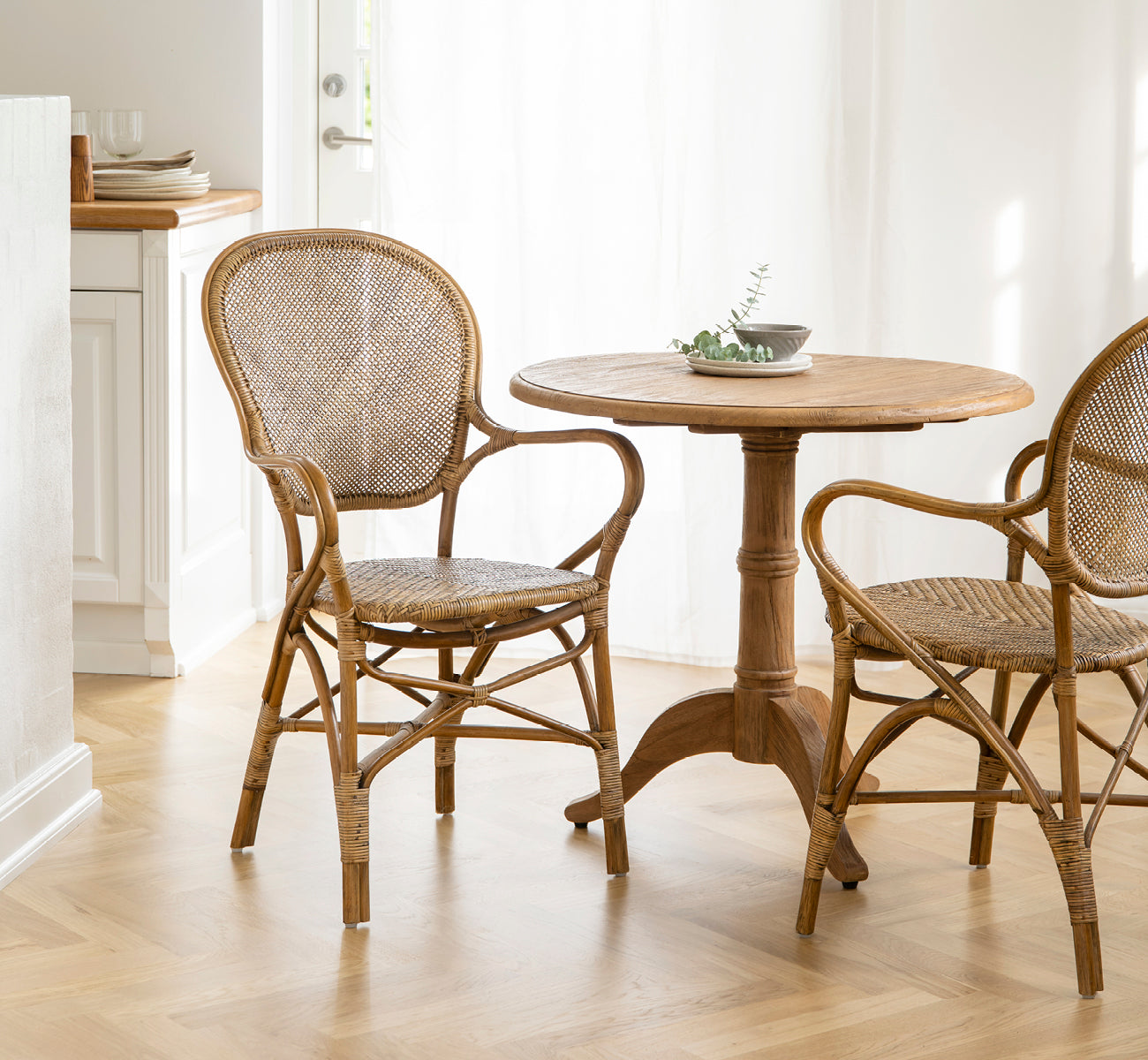Dies ist ein Lifestyle-Bild von Rossini Esszimmer-Armlehnstuhl in antique rattan. Kombiniert mit einem runden Cafétisch in einem hellen, luftigen skandinavischen Interieur.