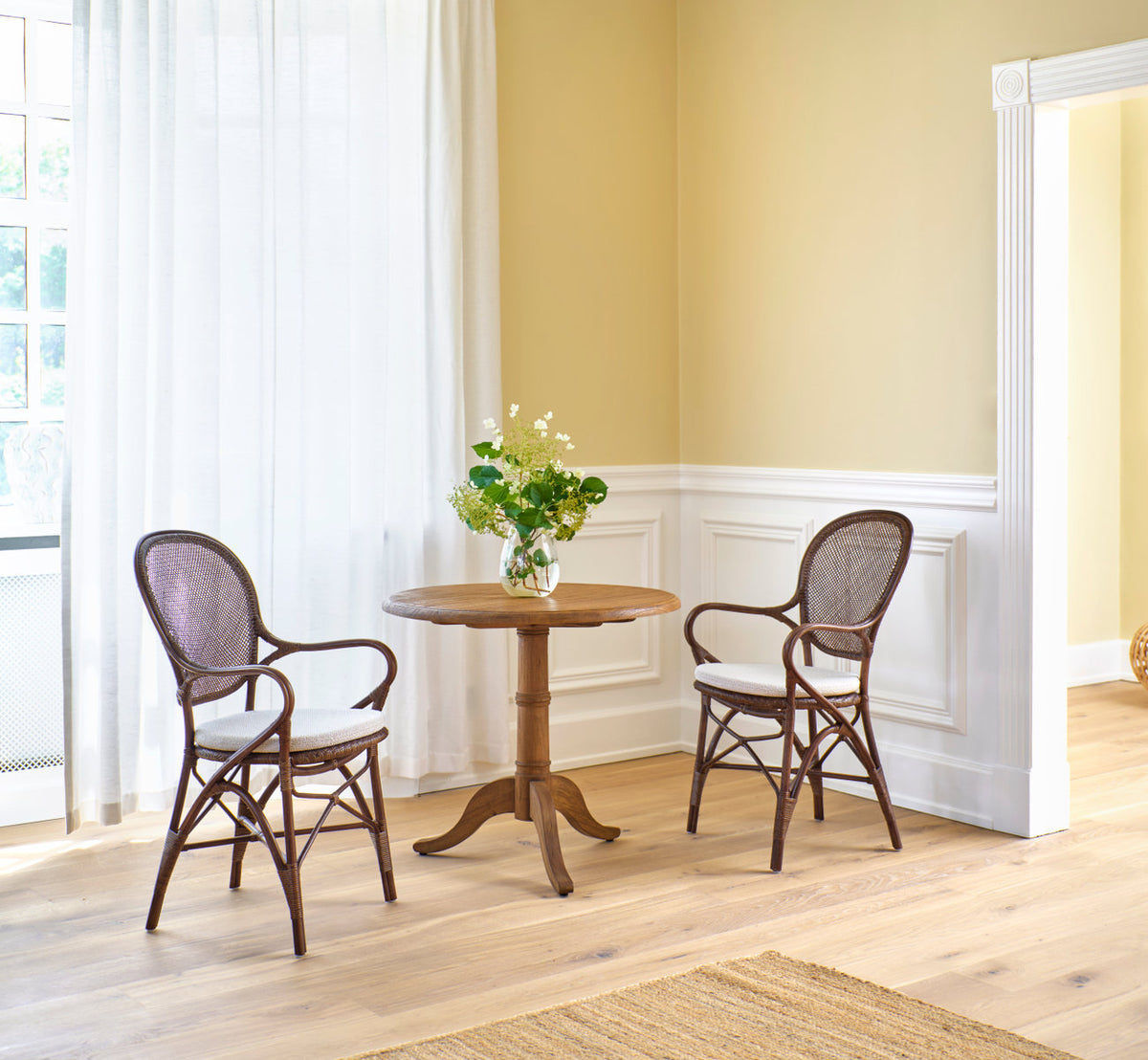 Dies ist ein Lifestyle-Bild von Rossini Esszimmer-Armlehnstuhl in brown rattan. Paar an einem Teak-Cafétisch vor einer weiß vertäfelten Wand.