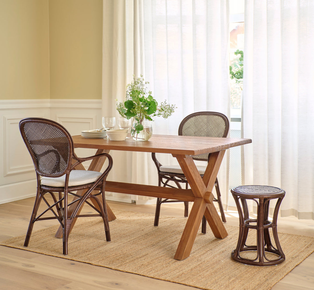 Dies ist ein Lifestyle-Bild von Rossini Esszimmer-Armlehnstuhl in brown rattan. Teaktisch, Rattanhocker und Juteteppich in einem hellen Raum.