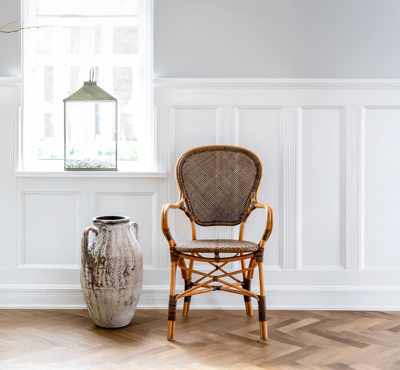 Dies ist ein Lifestyle-Bild von Rossini Esszimmer-Armlehnstuhl in cherry rattan. In einem vertäfelten Raum mit Fischgrätboden neben einer rustikalen Urne aufgestellt.