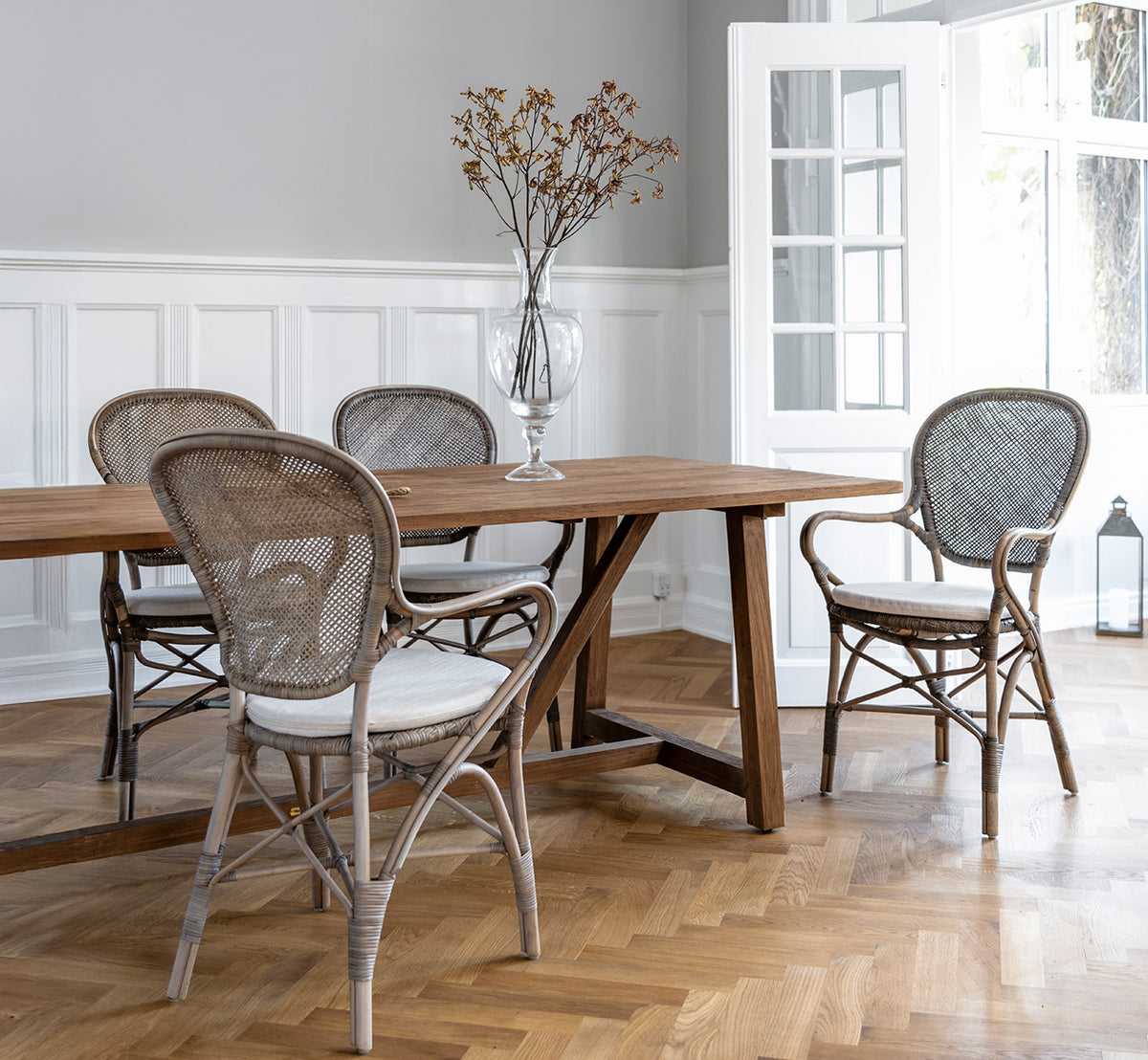 Dies ist ein Lifestyle-Bild von Rossini Esszimmer-Armlehnstuhl in taupe rattan. Um einen Holzesstisch auf einem Fischgrätparkettboden.