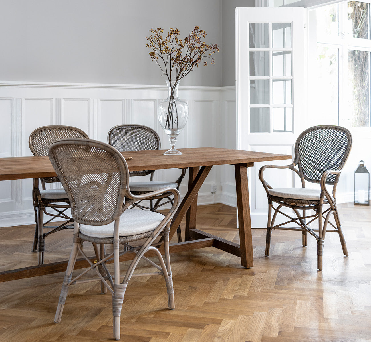 Dies ist ein Lifestyle-Bild von Rossini Esszimmer-Armlehnstuhl in taupe rattan. Um einen Holzesstisch auf einem Fischgrätparkettboden.