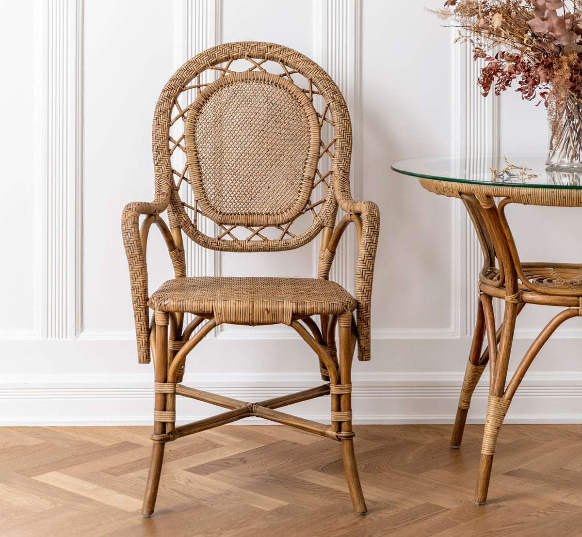 Dies ist ein Lifestyle-Bild von Romantica Esszimmerstuhl in antique rattan. Neben einem Glas-Rattantisch mit Trockenblumen in einem vertäfelten Raum platziert.