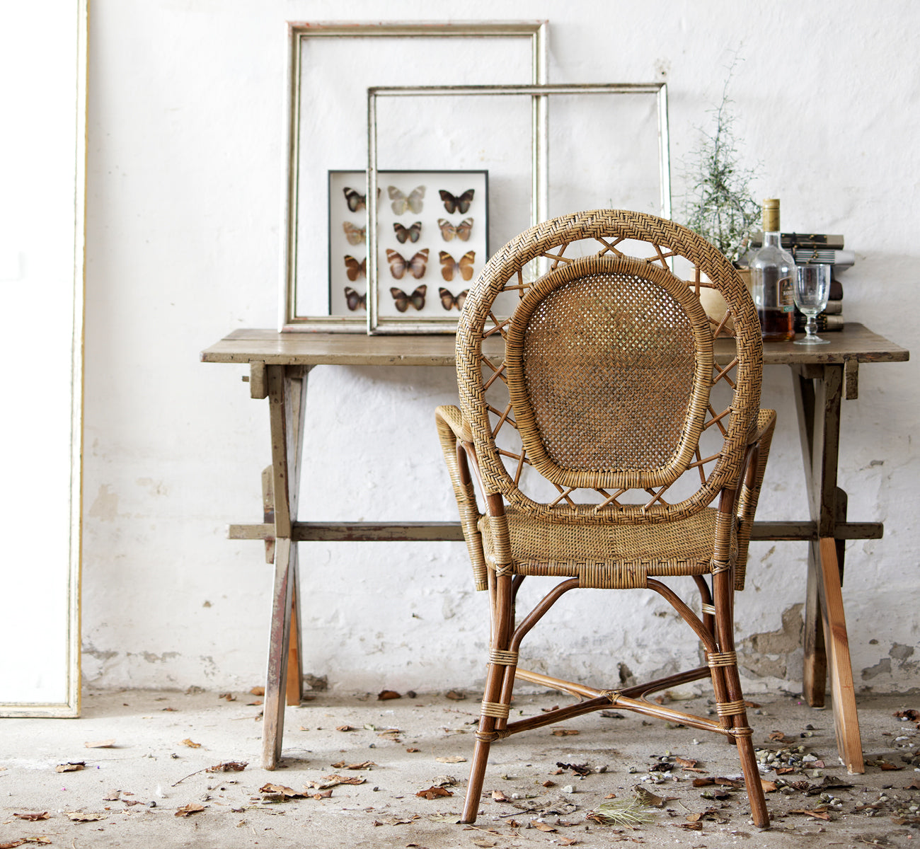 Dies ist ein Lifestyle-Bild von Romantica Esszimmerstuhl in antique rattan. An einem rustikalen Holztisch mit gerahmter Kunst in einem Vintage-Ambiente gestylt.