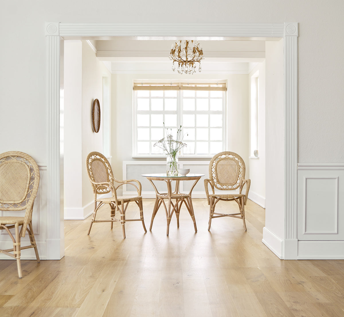 Dies ist ein Lifestyle-Bild von Romantica Esszimmerstuhl in natural rattan. Um einen runden Tisch in einem eleganten Raum mit Kronleuchter und weißer Vertäfelung.