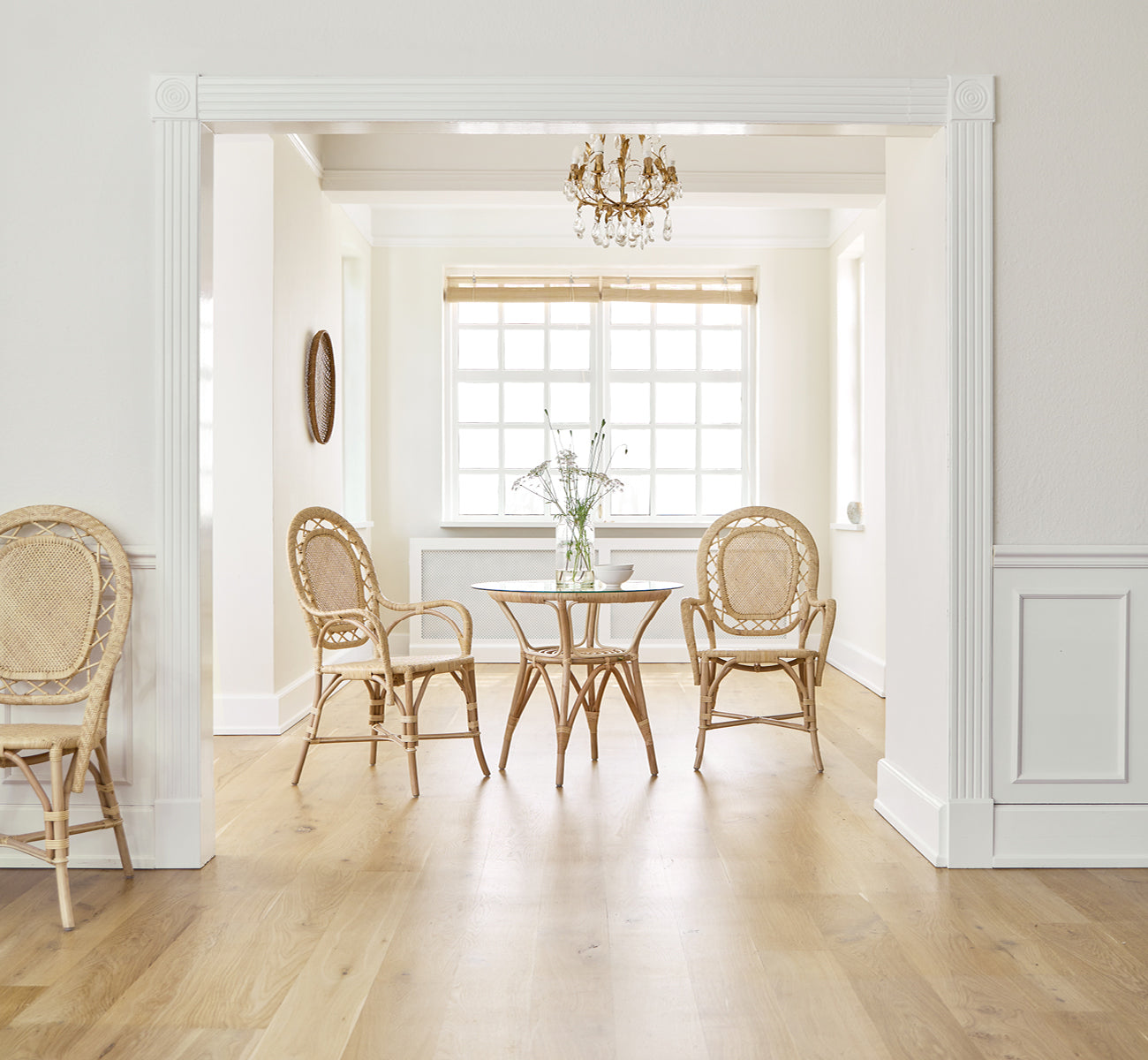 Dies ist ein Lifestyle-Bild von Romantica Esszimmerstuhl in natural rattan. Um einen runden Tisch in einem eleganten Raum mit Kronleuchter und weißer Vertäfelung.