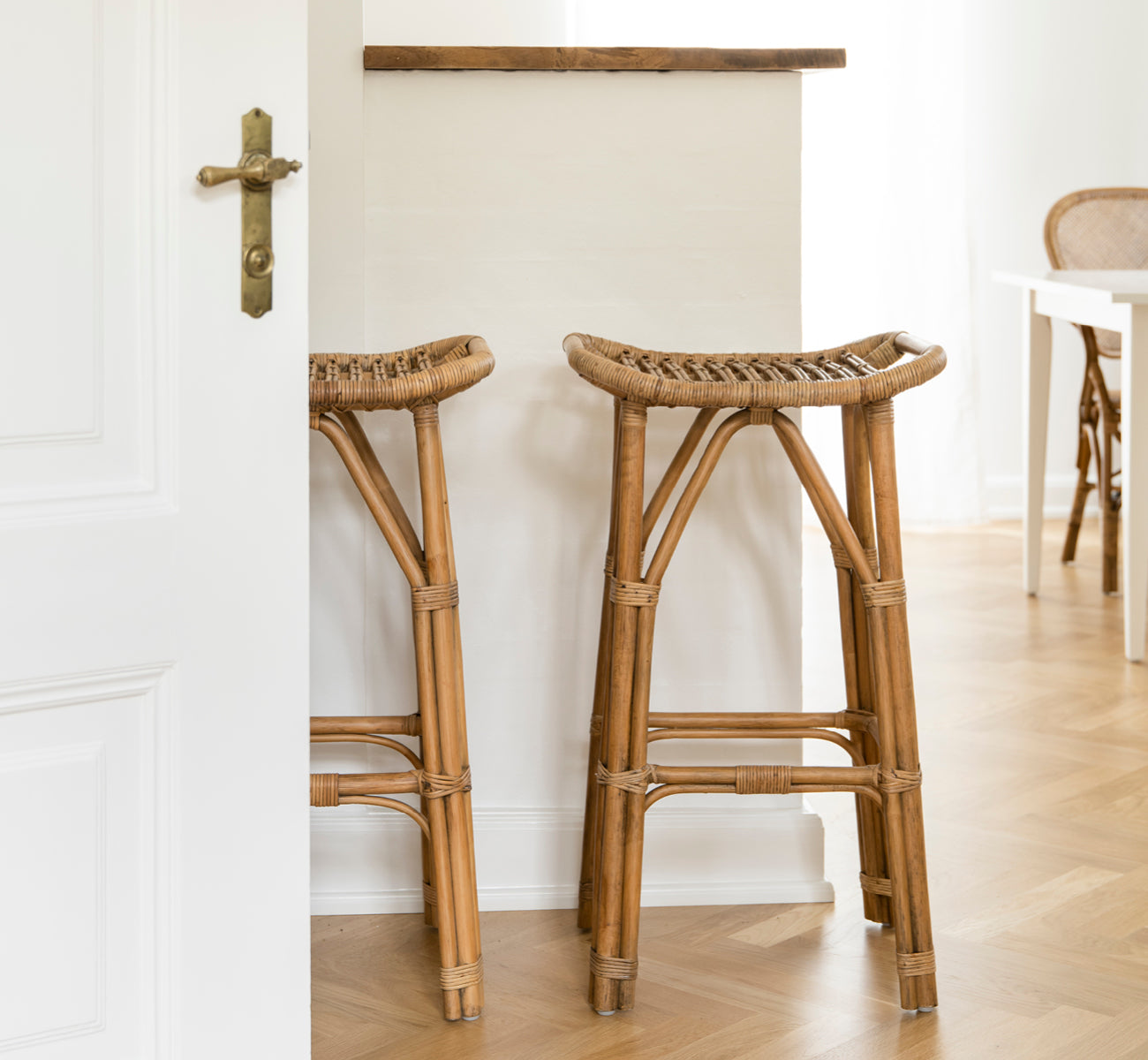 Dies ist ein Lifestyle-Bild von Salsa Barhocker in antique rattan. Ein Paar an einer weißen Wand auf Fischgrätparkett platziert.