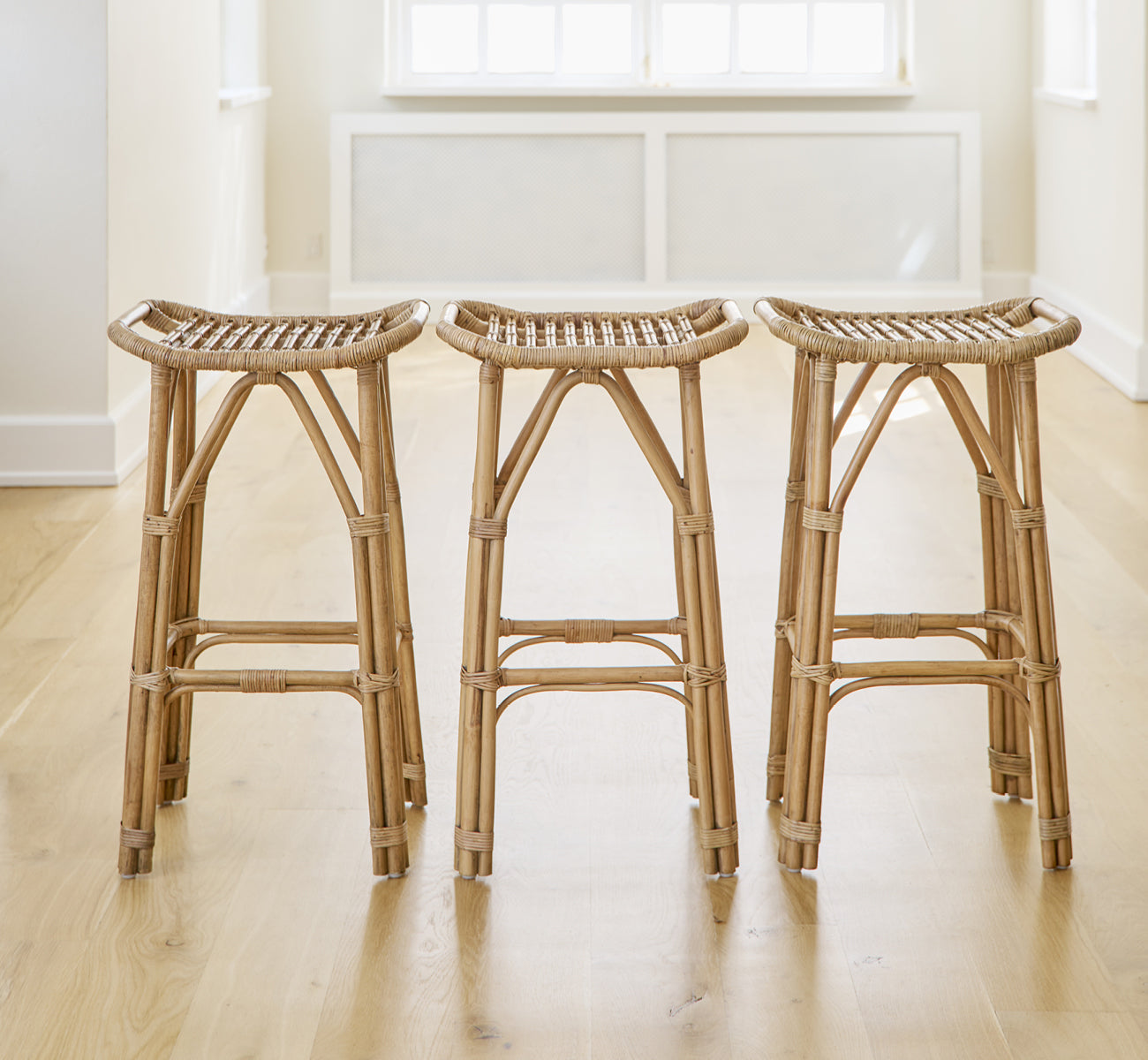 Dies ist ein Lifestyle-Bild von Salsa Barhocker in antique rattan. Ein Paar an einer weißen Wand auf Fischgrätparkett platziert.