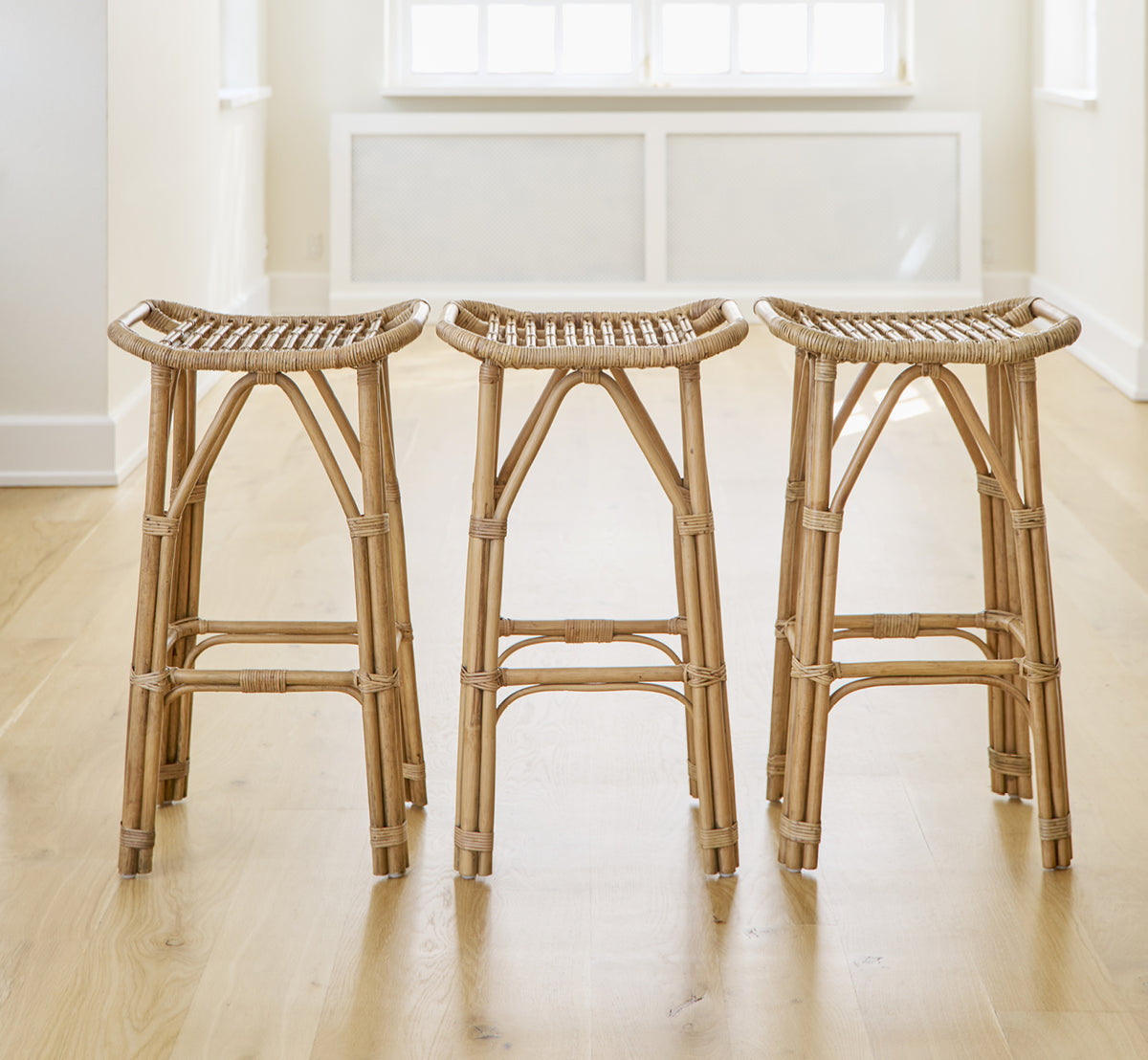 Dies ist ein Lifestyle-Bild von Salsa Barhocker in antique rattan. Ein Paar an einer weißen Wand auf Fischgrätparkett platziert.