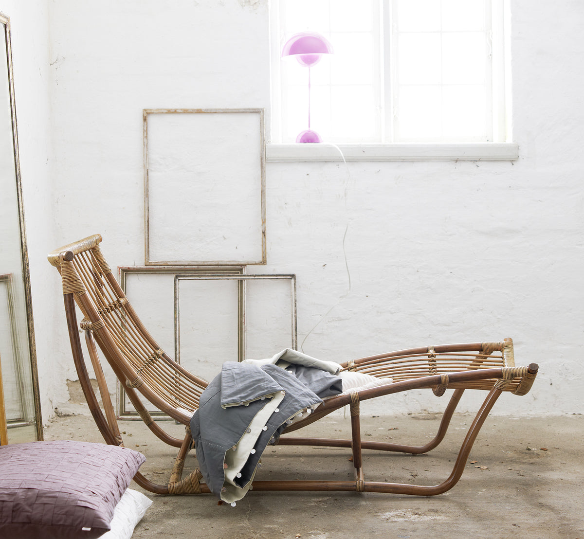 Dies ist ein Lifestyle-Bild von Michelangelo Daybed i antique rattan. Gestylt mit Leinendecken in einem weißen Studio mit Vintage-Rahmen.