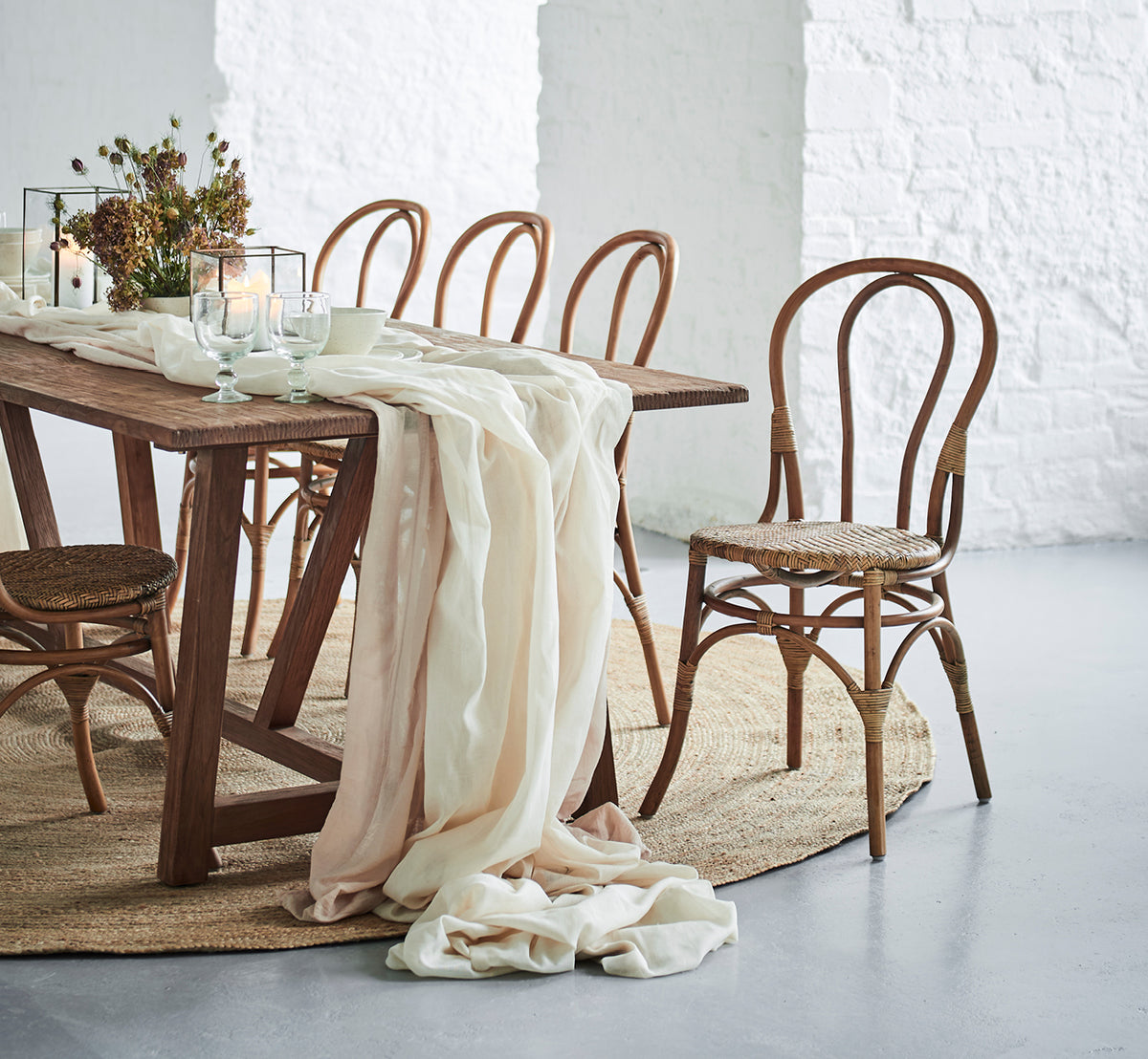 Dies ist ein Lifestyle-Bild von Lulu Esszimmerstuhl in antique rattan. An einem rustikalen Tisch mit Leinenstoff und Kerzen vor einer weißen Backsteinwand.