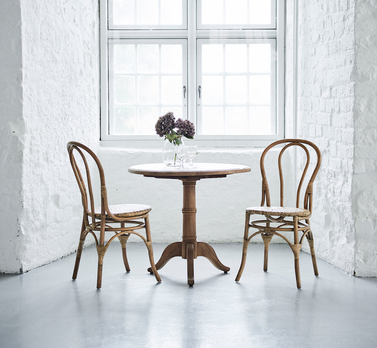 Dies ist ein Lifestyle-Bild von Lulu Esszimmerstuhl in antique rattan. Zwei Stühle flankieren einen runden Tisch mit Blumen in einem hellen weißen Backsteininterieur.
