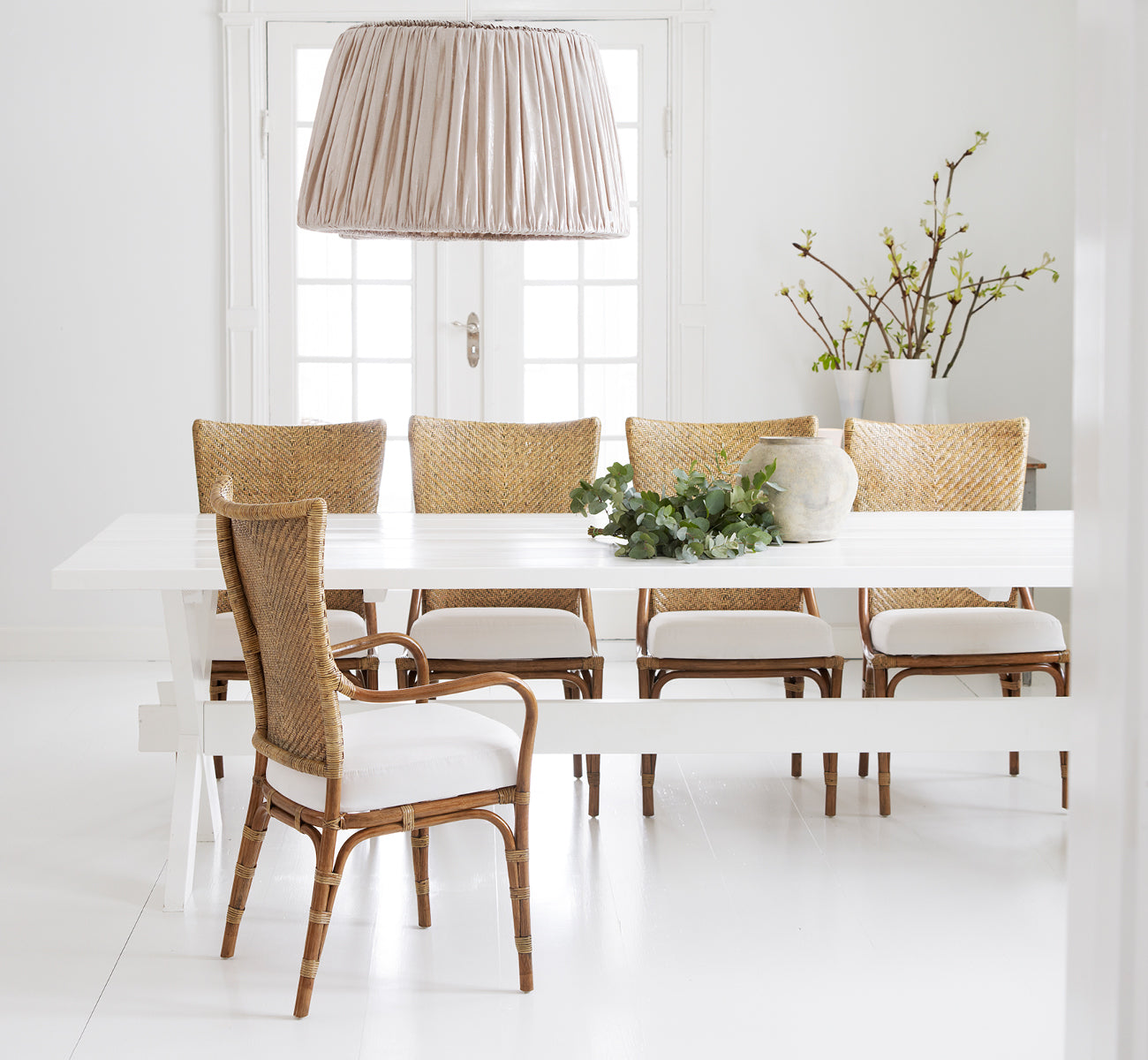 Dies ist ein Lifestyle-Bild von Melody Dining Armchair i antique rattan. Um einen weißen Tisch mit Leinen-Pendelleuchte und Frühlingszweigen.
