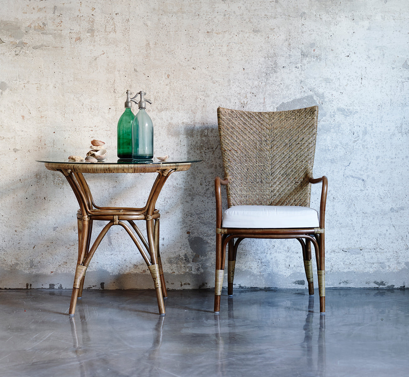 Dies ist ein Lifestyle-Bild von Melody Dining Armchair i antique rattan. Kombiniert mit einem Beistelltisch mit Glasplatte vor einer Betonwand.