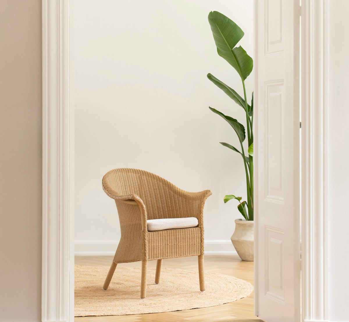 Dies ist ein Lifestyle-Bild von Classic Esszimmerstuhl in natural Loom Rattan Frame, in einem hellen Interieur mit weißen Wandpaneelen und.