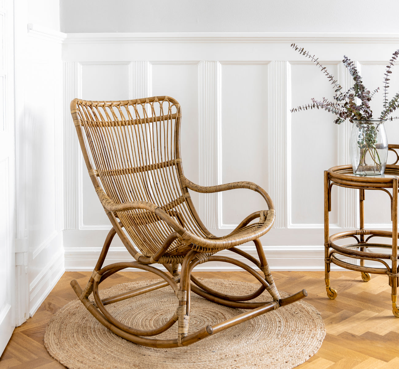 Dies ist ein Lifestyle-Bild von Monet Rocking Chair i antique rattan. In einem Raum mit Schmetterlings-Tapete und einem Rattan-Beistelltisch mit Glasplatte.