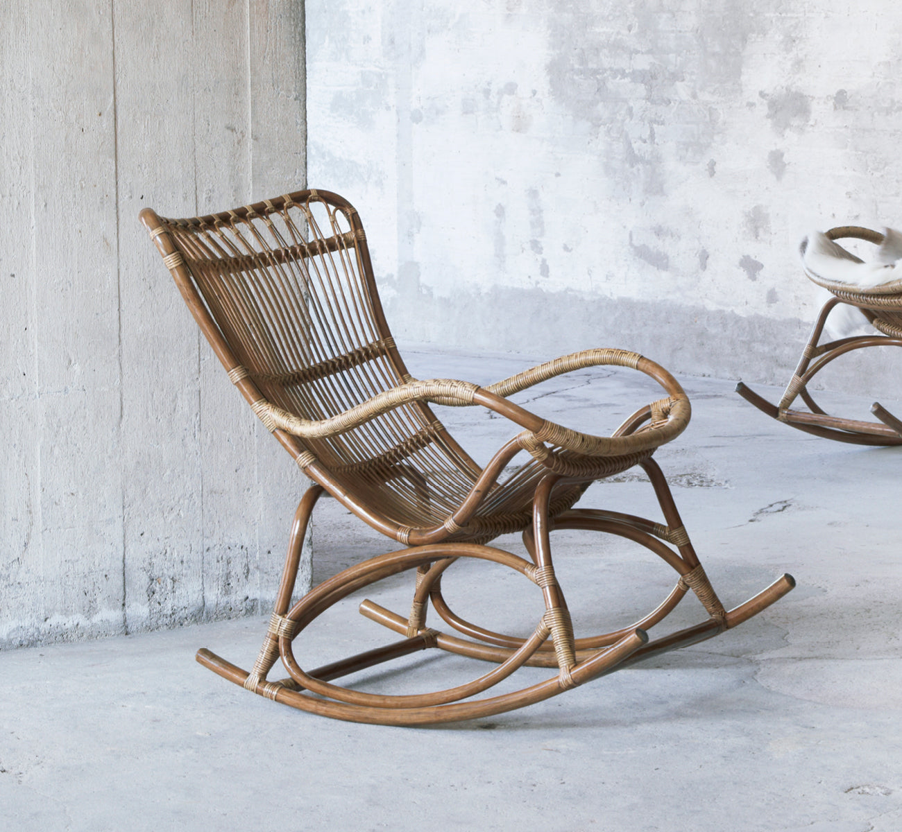 Dies ist ein Lifestyle-Bild von Monet Rocking Chair i antique rattan. In einem rustikalen Betonraum mit einem weiteren Schaukelstuhl im Hintergrund.