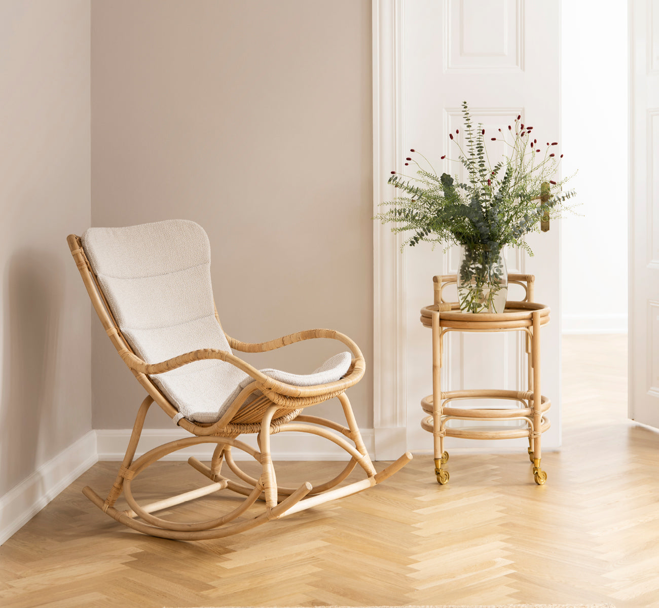 Dies ist ein Lifestyle-Bild von Monet Rocking Chair i natural rattan. In einem Raum mit rosafarbenen Wänden, Fischgrätparkett und Eukalyptus.