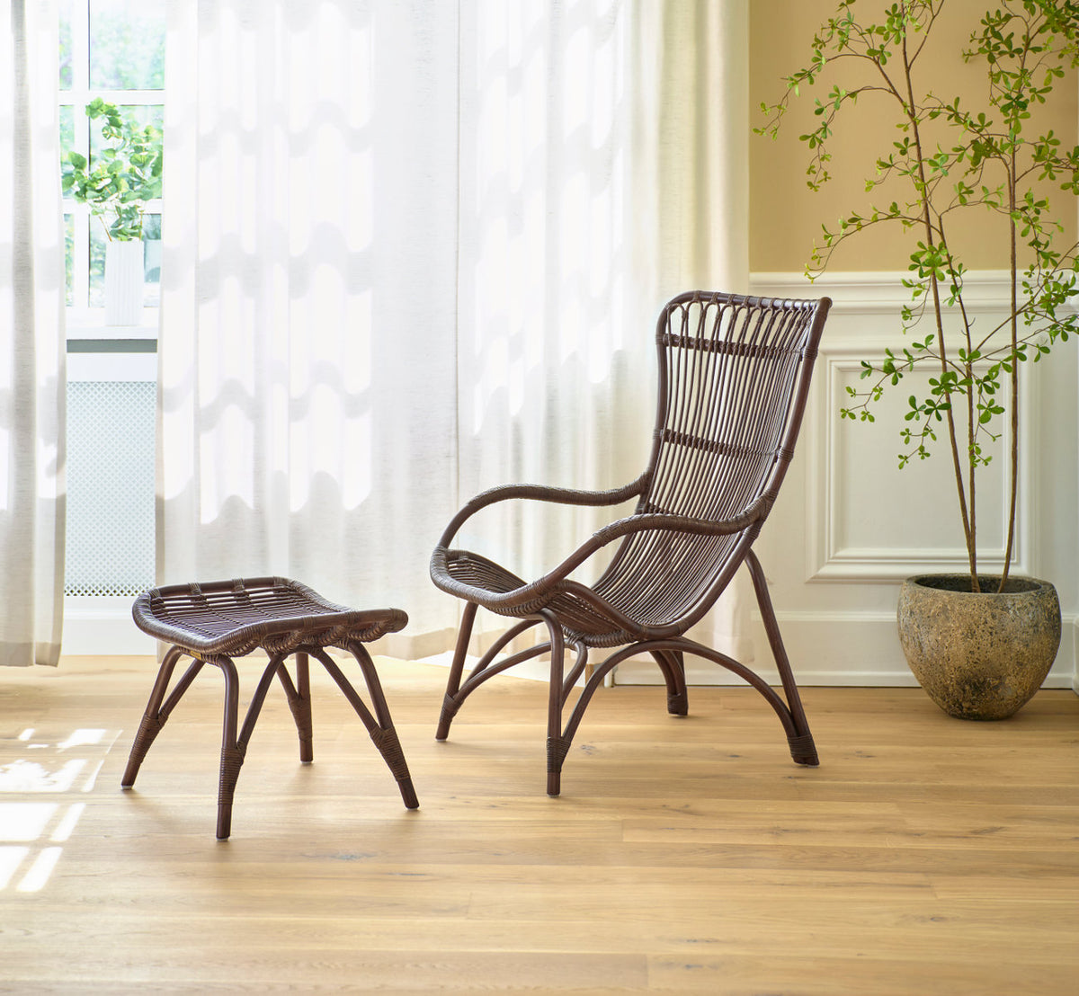 Dies ist ein Lifestyle-Bild von Monet Lounge-Sessel in brown rattan. Mit Fußhocker und Pflanze vor leichten Vorhängen.