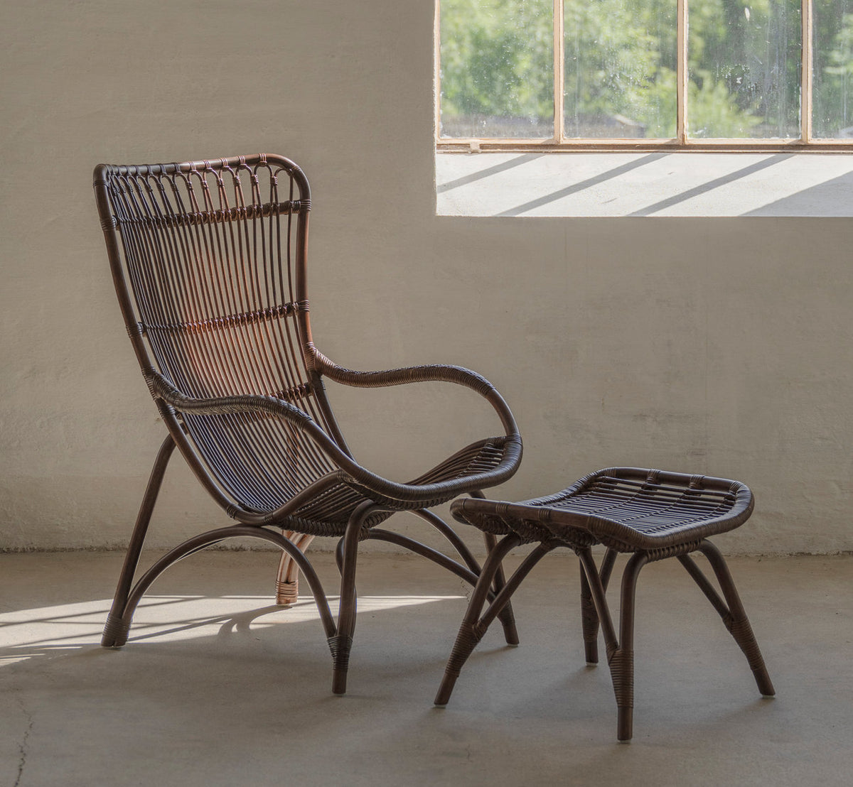 Dies ist ein Lifestyle-Bild von Monet Lounge-Sessel in brown rattan. Mit Fußhocker in einem sonnigen Raum mit Betonboden.