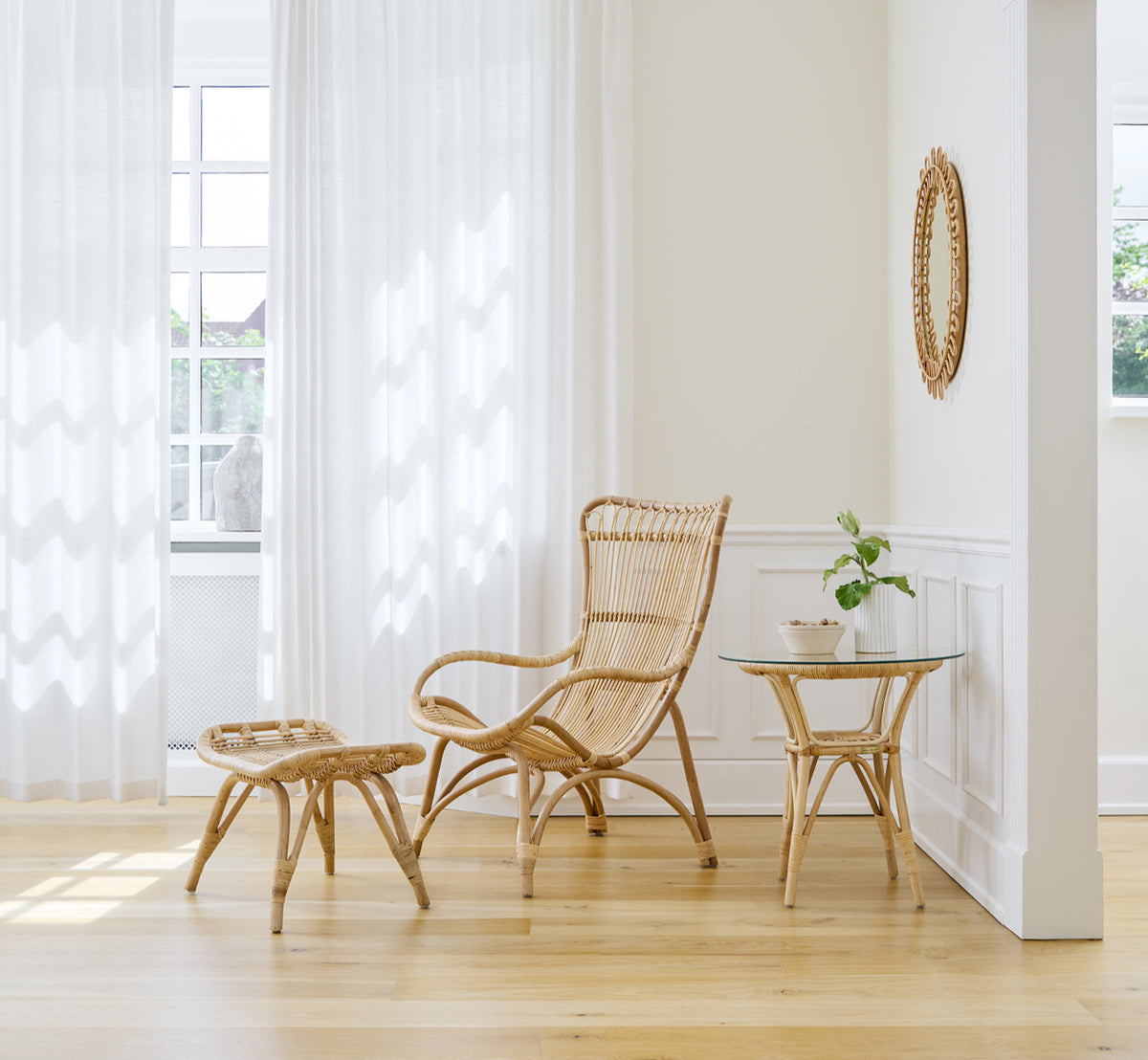 Dies ist ein Lifestyle-Bild von Monet Lounge Chair i natural rattan. Mit Fußhocker in einem hellen Raum mit Eichenboden und transparenten Vorhängen.