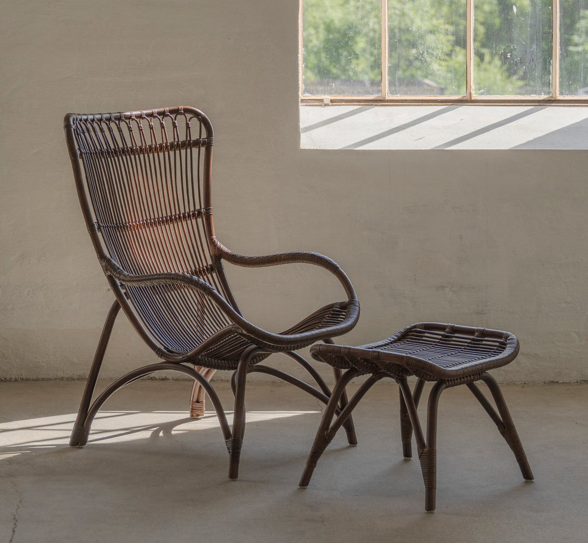 Dies ist ein Lifestyle-Bild von Monet Fußhocker in brown rattan. Mit Lounge-Sessel in einem sonnigen Raum mit Betonboden.