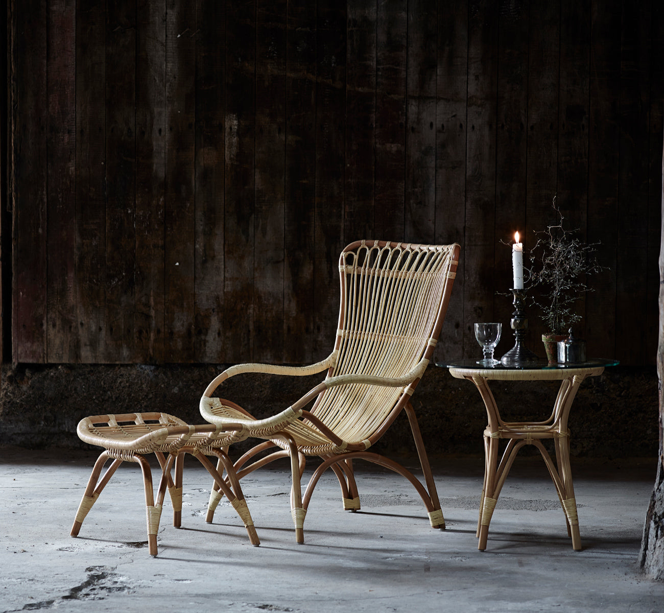 Dies ist ein Lifestyle-Bild von Monet Footstool i natural rattan. Mit Lounge-Sessel in einem dunklen rustikalen Scheunen-Setting mit Glaswaren.