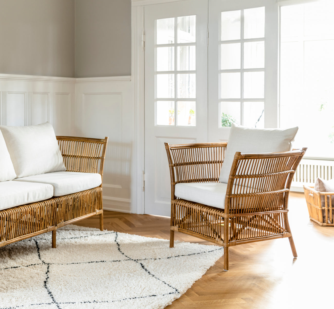 Dies ist ein Lifestyle-Bild von Donatello Lounge-Sessel in antique rattan, in einem hellen Wohnzimmer mit Fischgrätparkett und üppigem Grün.