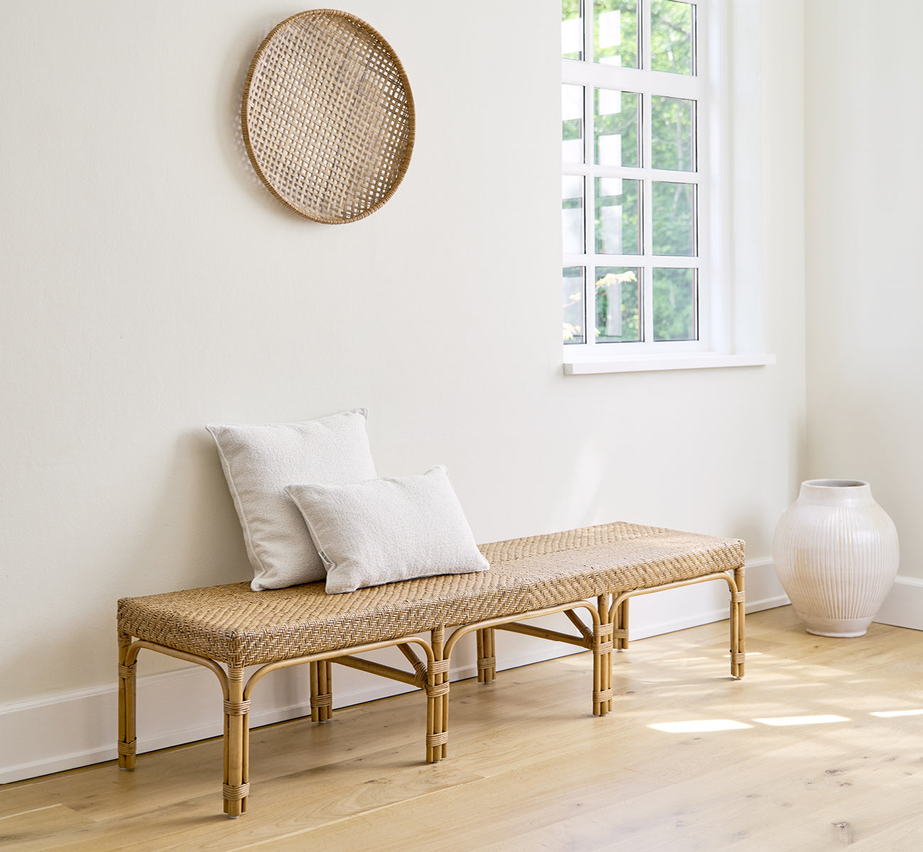Dies ist ein Lifestyle-Bild von Luis Bank in antique rattan. Mit Leinenkissen in einem hellen weißen Raum mit Wanddeko und weißer Vase gestylt.